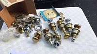 Top view of assorted door knobs mostly brass finish plus some chrome, with mounting plates and screws on white tablecloth.