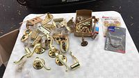 Varied brass and copper finish door hardware arranged on a white tablecloth including lever handles, knobs, hinges, latch plates, and some packaged items.