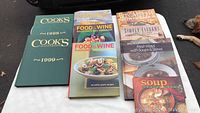 Photo showing nine cookbooks and magazines arranged on a white surface. Includes two Cook's Illustrated hardcover books 1998 and 1999 editions, two Food & Wine magazines, Box Appetit, Simply Elegant, Fresh & Warmer with Soups & Stews, Soup cookbook, and Cooking Annual Recipes 2000.