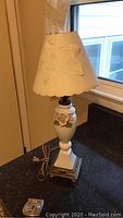 Front view of vintage small cream porcelain floral table lamp on black counter near window, showing base, shade, and power cord with plug.