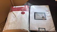 Two off white tablecloths shown: one packaged Huntington Home Holiday set with 6 napkins, and one packaged Rosedale spill proof tablecloth.
