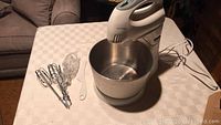 Photo showing white electric stand mixer attached to stainless steel bowl with beaters and accessories to the side on table.