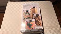 Closed unopened box of SelectHome set of 4 glass canisters shown on beige patterned tablecloth.