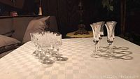 Wide view showing both sets of cordial glasses arranged on a checkered tablecloth, set of 6 faceted glasses on left, set of 8 smooth silver-accented glasses on right.