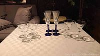 Photo showing 4 wide champagne glasses on left, 4 wine glasses with dark blue stems and bases in center, 2 martini glasses on right, arranged on a checkered white tablecloth.