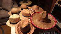 Seven straw hats arranged on a white surface in a living room, showing different styles including 4 Mexican sombrero hats with wide brims, one trimmed in red and white, and three other woven straw hats.