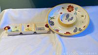 Photo showing both the 14-inch round chip and dip serving tray with butterfly and floral design and the 14-inch rectangular divided serving tray with three compartments and ladybug corner motifs on a white fabric background.