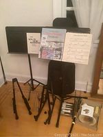Two adjustable black metal music stands with sheet music and a soft guitar case standing upright near them, with additional items on floor including drumsticks and harmonica.
