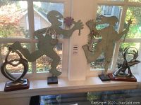 Wide view of four decorative dancing figures arranged on a white windowsill in front of a window with autumn leaves visible outside. Two large metal silhouette dancing figures hold colorful hanging accessories and smaller painted figures.