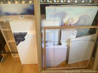 Multiple wrapped and loose canvases leaning against a wall and a gold-tone frame