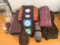 Multiple folded and rolled yoga mats in purple, orange patterned mat, gray rolled mat, with small decorative items in front including a dark goddess plaque, white candles and other small accessories.