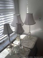 Four bedside lamps with glass bases and white fabric shades placed on marble-topped tables near window blinds.
