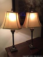 Pair of lit table lamps on wooden surface with beige square bell-shaped shades and metal bases with carved ornamentation.
