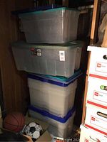 Stack of six Rubbermaid storage containers with green and blue lids, stored in a wooden shelving space with a basketball and soccer ball on the floor.