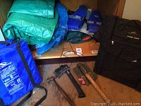 Photo of folded tarps in blue and green, blue floatation cushions, two Sterns blue life jackets, and black Lounge Lizard carrying bag on shelf and floor.