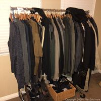 Full view of multiple men's sweaters, jackets, and trench coats hanging on a metal clothing rack on wooden hangers with baskets of socks underneath.