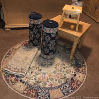Photo showing the round floral-pattern rug on the floor, the two rolled black runners, the beige runner folded on the rug, with a wooden side table and a small wooden step stool placed on the side table.
