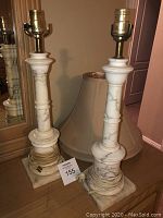 Full view of two vintage marble lamps showing white marble bodies with unique veining and brass sockets, placed on a wooden surface in front of a mirror. Power cords wrapped around the base.