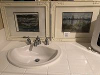 Two white-framed pictures placed beside a sink, showing embossed corner details; one picture depicts water lilies on a pond and the other a skyline reflected on water.