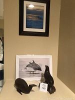 Overall view showing two hand carved wooden animal figurines (whale and seal) and two framed photos (dolphin and sea scene) arranged against a wall.