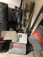 Full view of Kirby G4 vacuum, Dirt Devil handheld vacuum, Kirby accessory boxes, and cleaning liquid container inside a storage area.