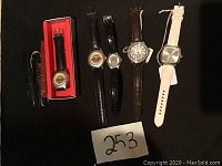 Five wristwatches laid out in a row on black fabric background with a '253' lot number card visible. Shows the watches' faces and straps from a front angle.
