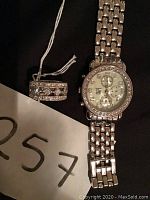 Photo showing silver-tone quartz watch with linked metal band and round face, and palladium-tone rhinestone ring with multiple rows of stones.