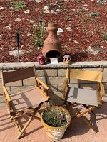 Wide shot showing two brown wooden director chairs with fabric seats and backs, a terracotta Takata pot on a stone wall, two colorful Day of the Dead skull decorations beside the pot, and a decorative plant in a pot between the chairs.