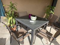 Full view of patio table and four matching chairs arranged around it, along with several potted plants on concrete patio surface.