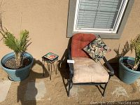 Photo showing the patio chair with beige seat cushion and burnt orange back cushion alongside the small tiled plant stand and two large potted palm plants.