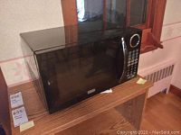 Front and side view of black Sunbeam microwave oven on wooden shelf with visible window reflection.