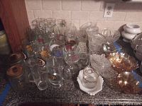 Photo shows multiple clear glass beer mugs with logos, cut glass bowls, some amber glass bowls, white ceramic bowls stacked, and jars on kitchen counter.