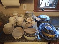Overview of Liberty Blue and Noritake dishes stacked on kitchen counter showing plates, bowls, cups, saucers, serving platters and other pieces.