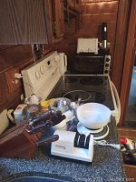 Photo showing a wooden knife block with several knives, a white Chefs Choice cutter sharpener, measuring cups, and a cooking scale with a white bowl on top, placed on a kitchen counter next to a stove.