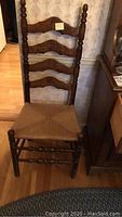 Frontal view of wooden chair with high ladder back and thatched woven seat on wood floor beside a cabinet.