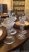 Photo showing assorted clear glass candle holders with floral etched designs on a wooden table, in front of decorative plates and other household items.
