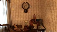 View of wall clock hanging, small table with baskets, crock, and trivets under it, showing arrangement of the lot