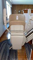 Front view of beige padded seat and backrest of stairlift mounted on rail along staircase. Shows armrests and footrest clearly, with visible wear and stains on fabric.
