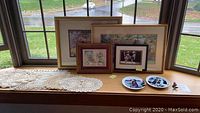 Window ledge with four framed pieces of art, two decorative plates, and multiple crocheted doilies.