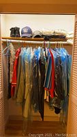 Closet rod with assorted suits, sport coats, jackets, and tops hanging on hangers. Two pairs of dark Baretrap boots, several ball caps, and folded sweaters are on upper shelf.