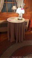 Photo of a round side table covered with fabric and lace cloth, with a white glass shade touch lamp placed on top, situated near a radiator and window.