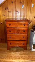Front view of wooden chest of drawers showing four drawers and round wooden knobs, against a wood-paneled wall and wooden floor.