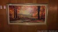 Framed wall art with a detailed autumn landscape showing fall-colored trees and a river, hung on a wood-paneled wall.