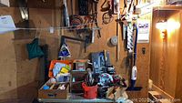 Wide view of wooden box with assorted hand tools, vintage saws hanging on the wall, some power tools on the table, and various small items in the lot.