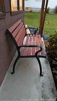 Full side view of the wood slat bench with black metal sides and armrests on a concrete porch with lawn background.