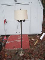 Full view of the tall antique floor lamp with cream fabric shade, showing weighted base and height outdoors.
