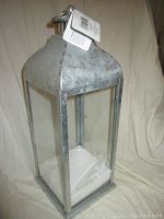 Front angled view of entire vented Christmas lantern showing galvanized metal top with vent, clear glass sides, square base, and attached manufacturer tags.