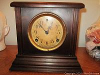 Front view of antique Sessions mantle clock, showing wooden case, clock face with brass bezel and dial with Arabic numbers. Missing front glass visible.
