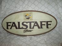 Image showing front view of an oval Falstaff Beer advertising sign with Off-white background and brown trim, Falstaff logo and text in black and yellow.
