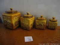 Front view of the four vintage mustard yellow square ceramic canisters with lids, embossed with Asian-influenced patterns.